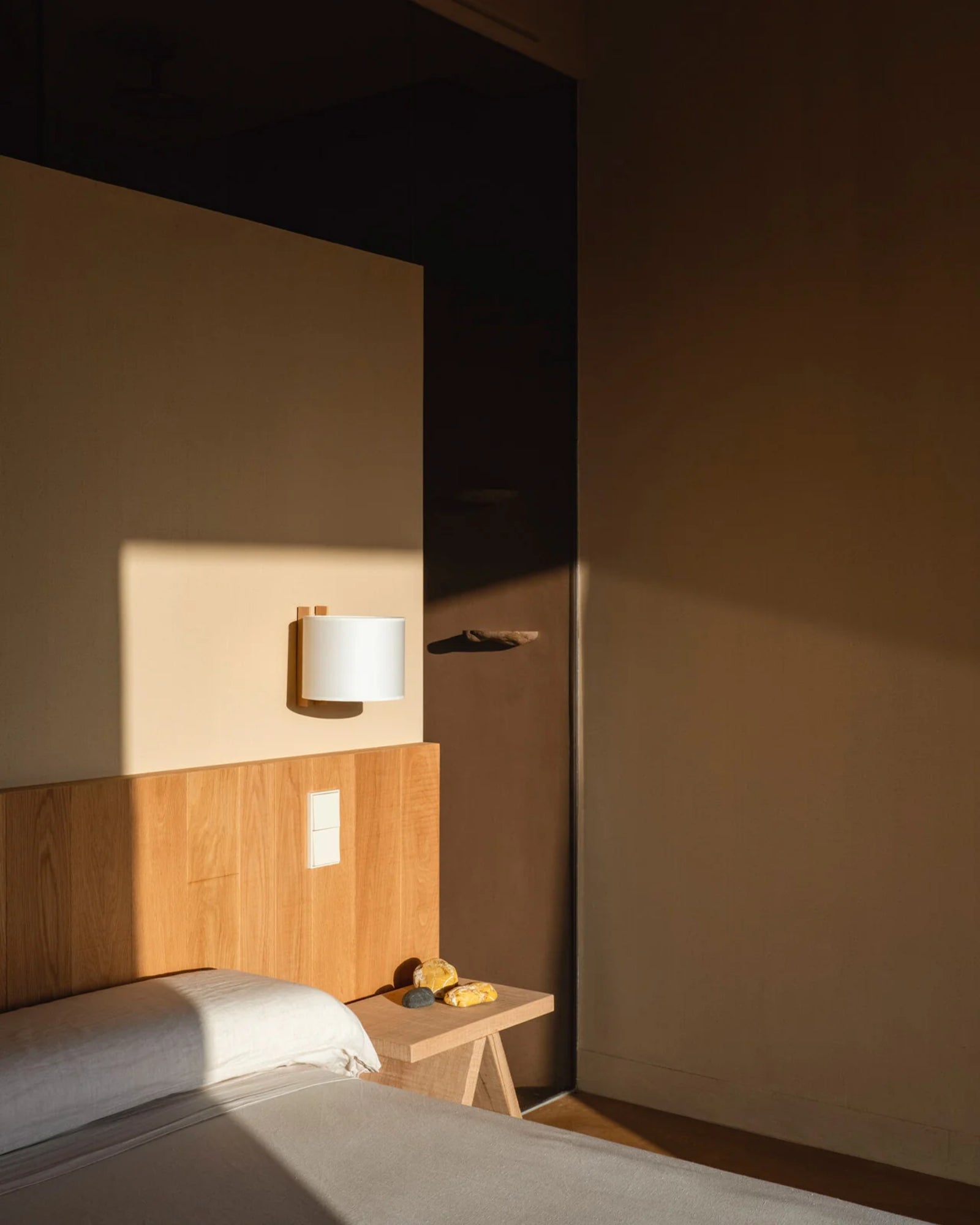 A minimalist TMM Corto wall light with a beige or white shade mounted on a wall, with a wooden channel in a bedroom setting.