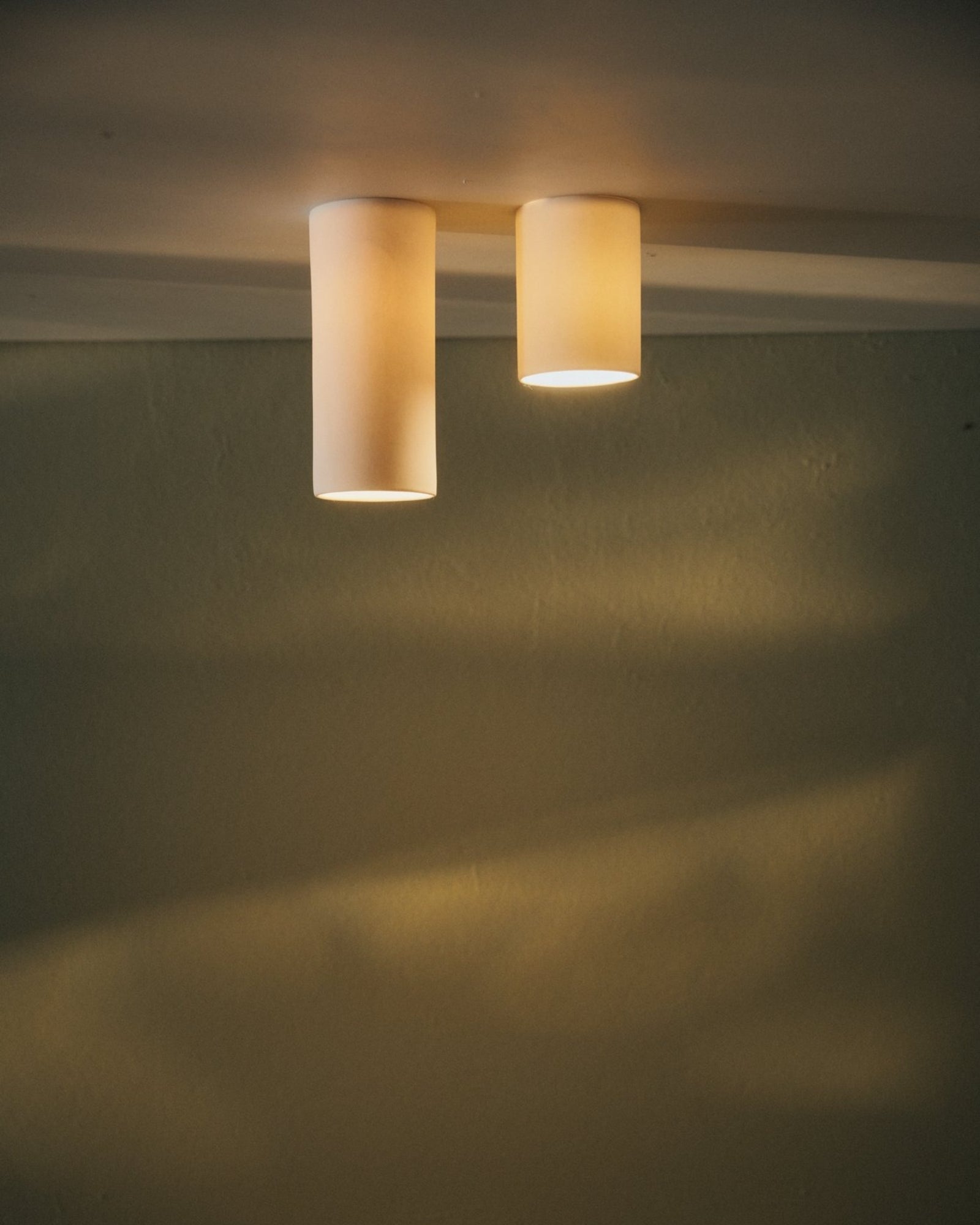 Two cylindrical ceiling lights called the Dusked Eos Ceiling Light by Studio Enti made of porcelain with a white finish, mounted on a ceiling and casting a soft glow on the wall.