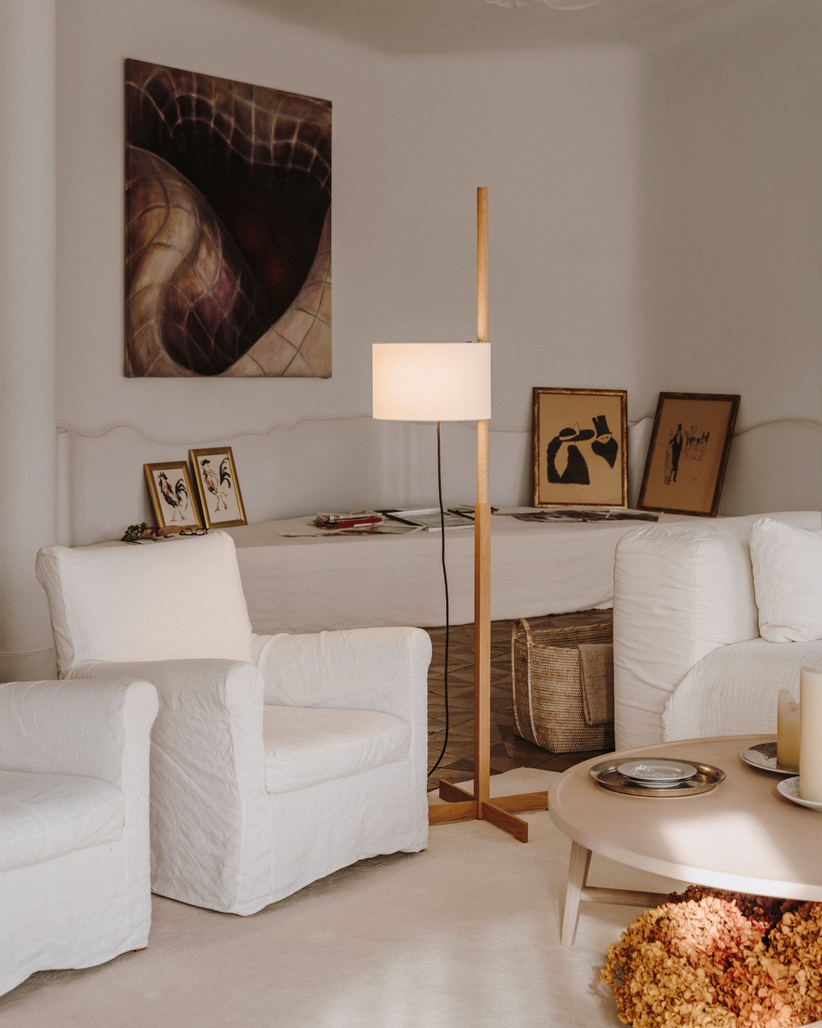 A TMM Floor Lamp with a beige shade, standing in a living room setting with a height-adjustable mechanism, flanked by two white armchairs and under a wall painting.