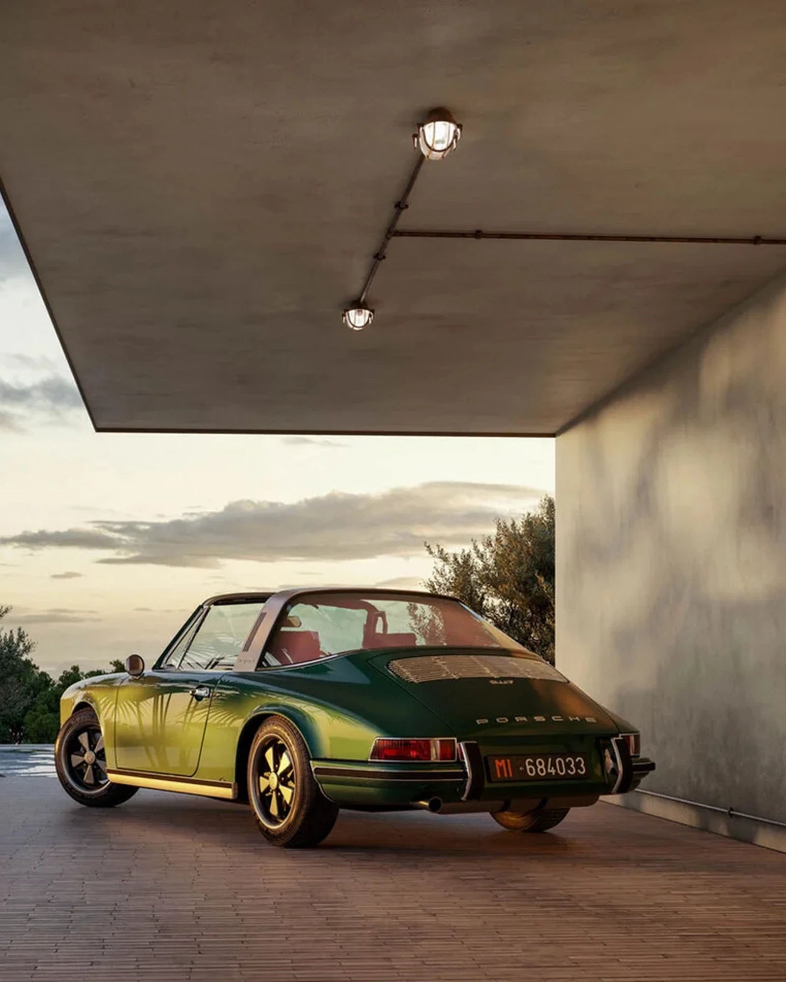 Green vintage car parked under a modern architectural structure with a sunset sky.