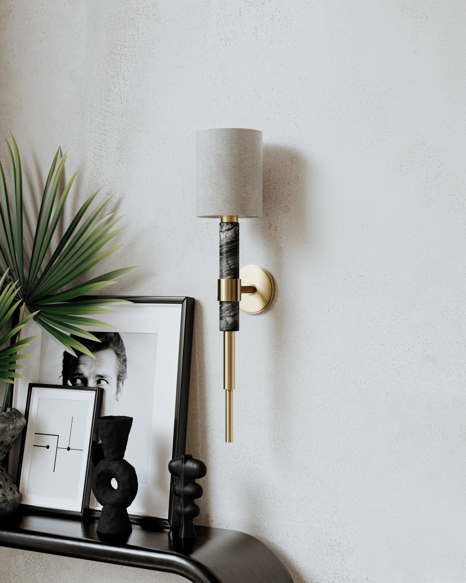 Decorative wall sconce with marble and gold details with a lampshade on a light gray wall, next to a framed picture and plant.